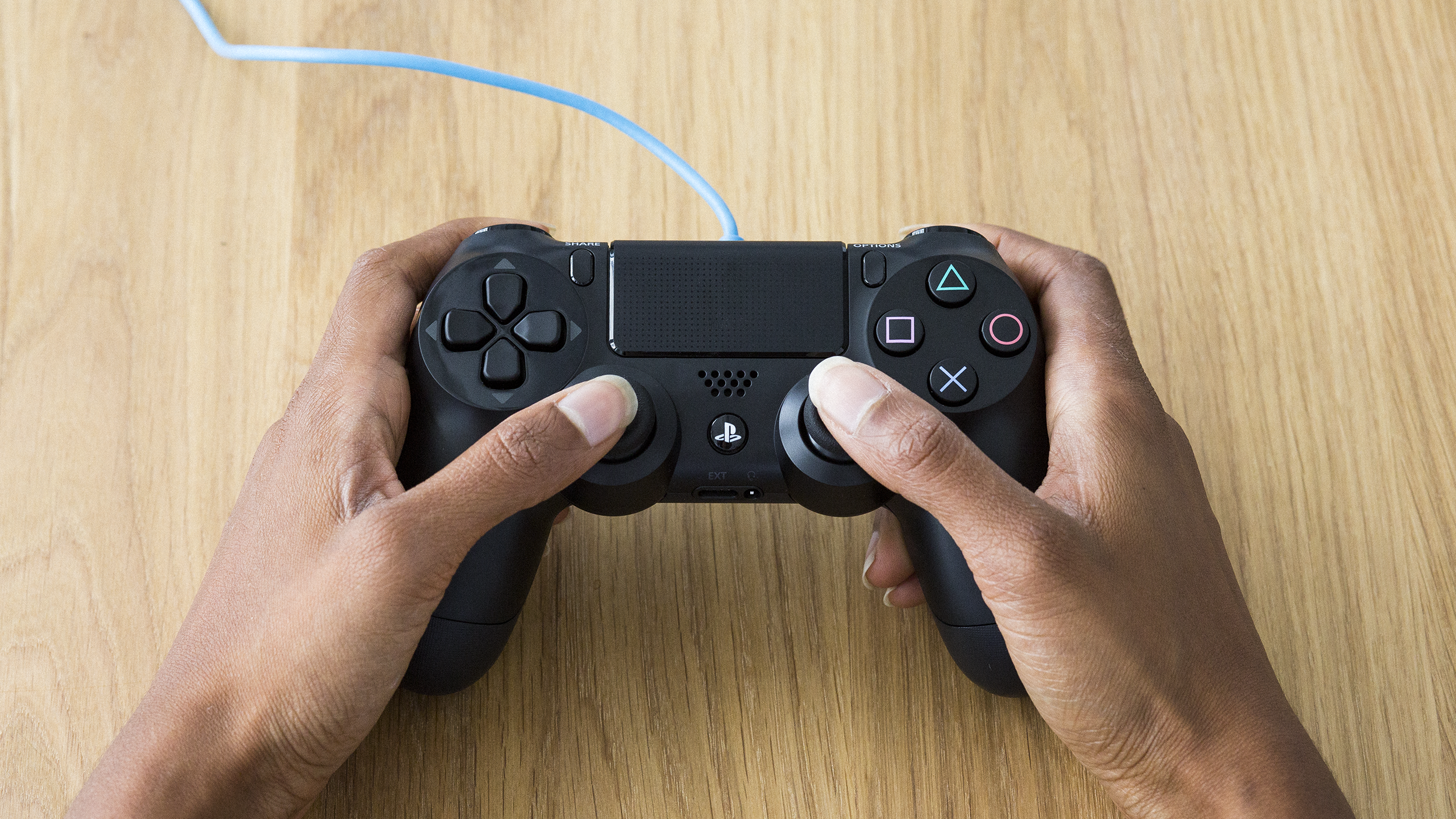 An overhead shot of two hands holding a wired Playstation 4 controller, over a wooden table.