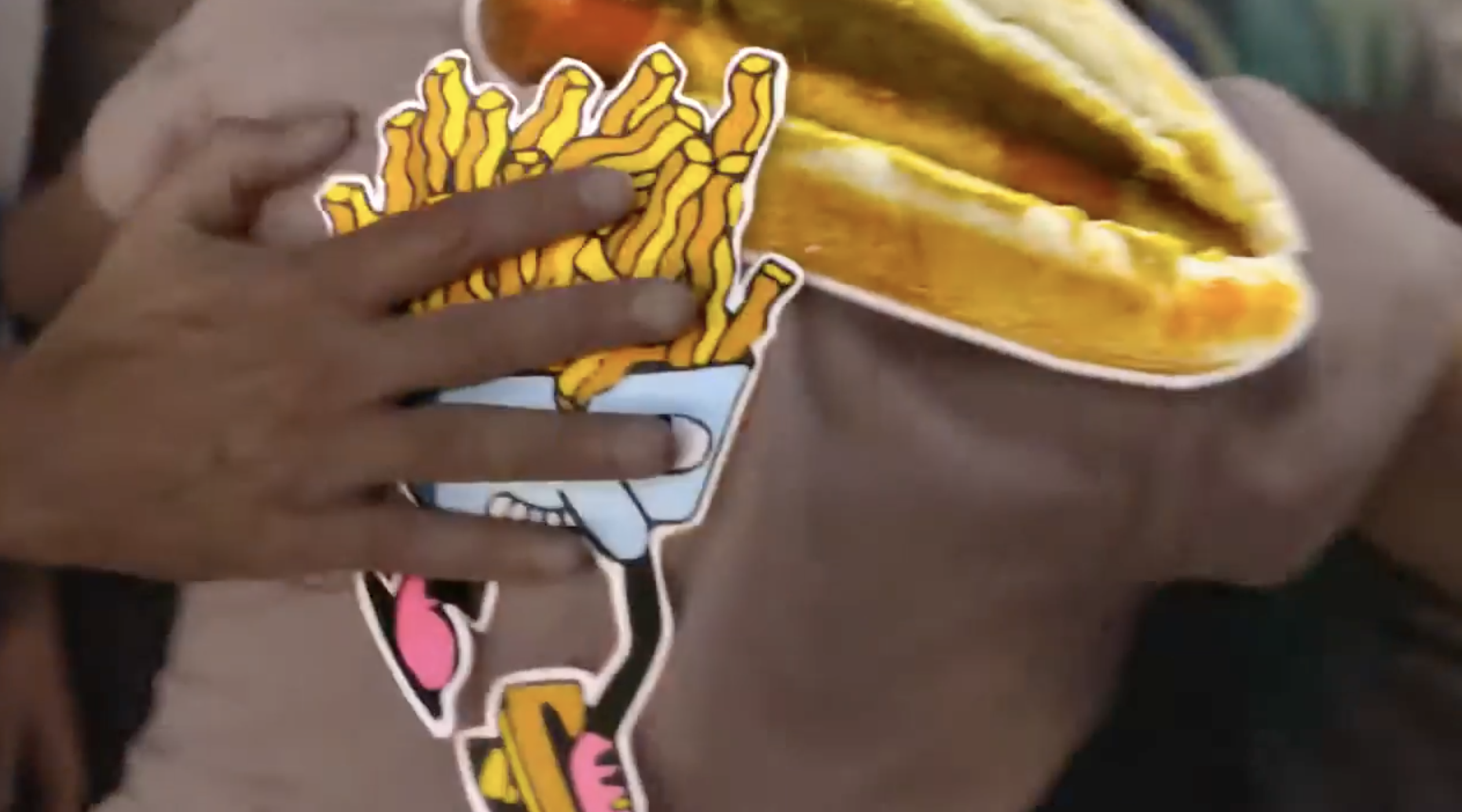A large sticker of a hot dog with mustard on the back of someone's shirt. A hand from offscreen is slapping another sticker onto it of an illustration of a box of French fries with a face and legs.