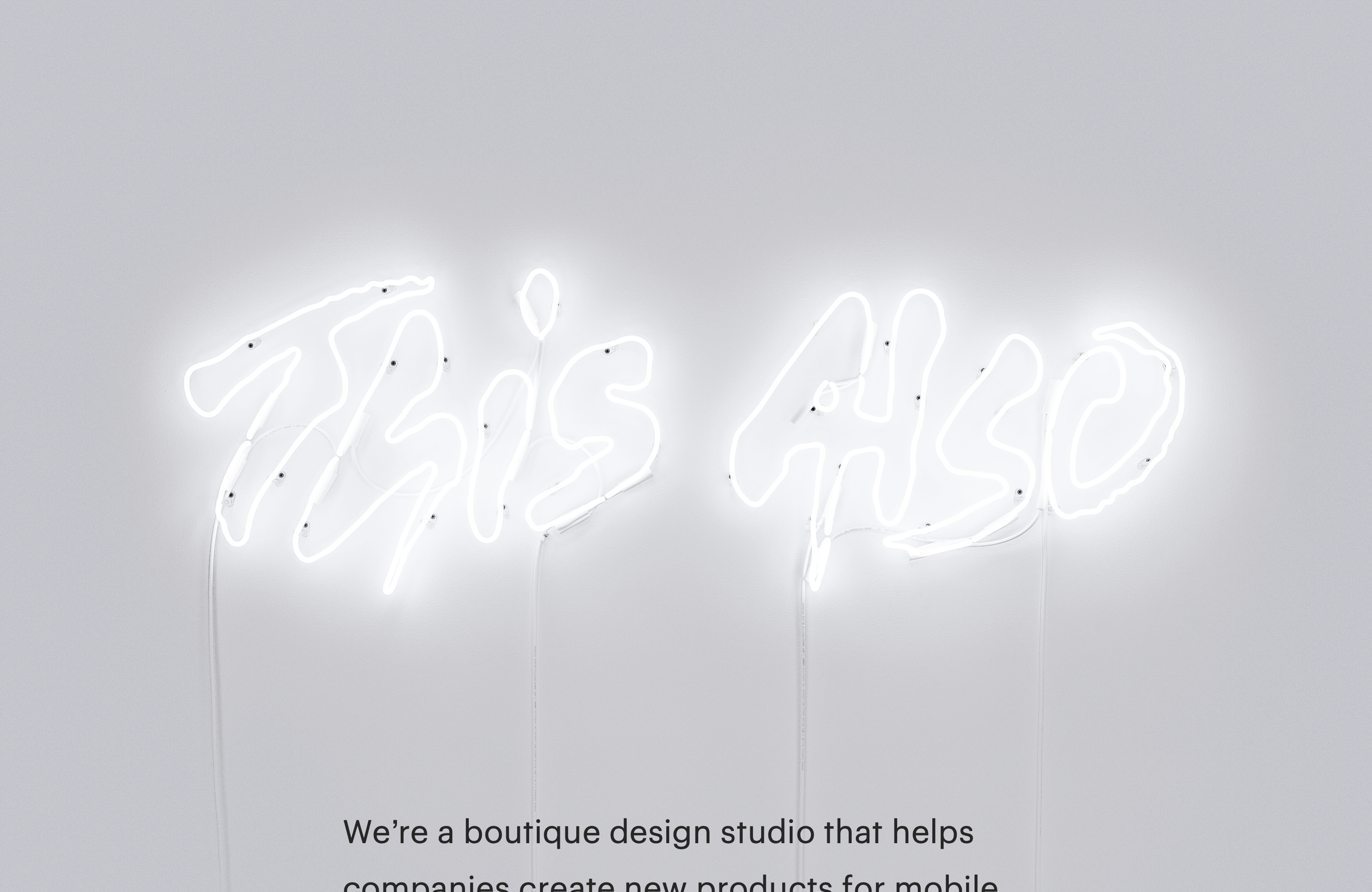 A lit up neon sign of the This Also logo on a light gray wall.