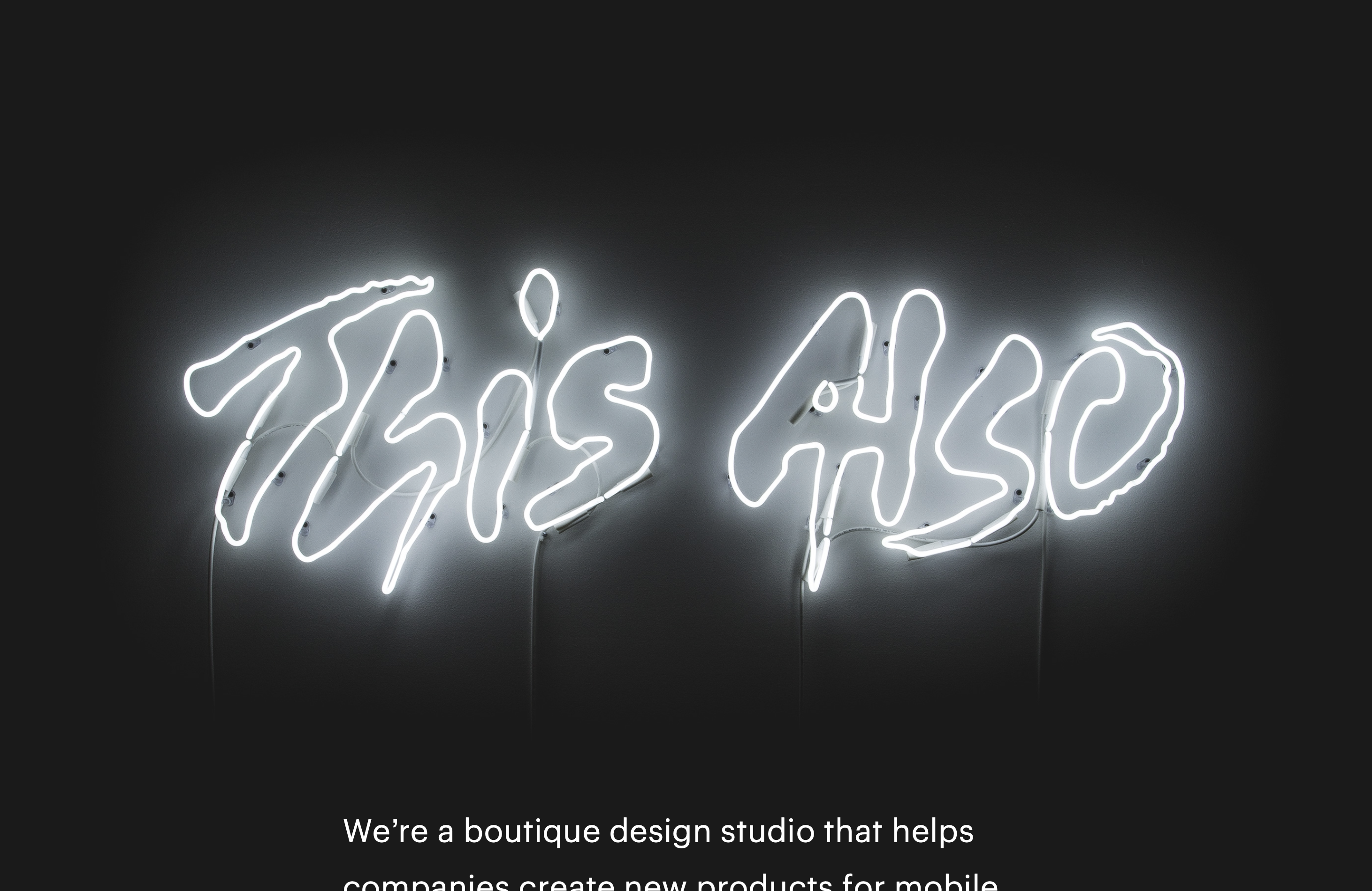 A lit up neon sign of the This Also logo in a dark room, faintly illuminating the white wall behind it.