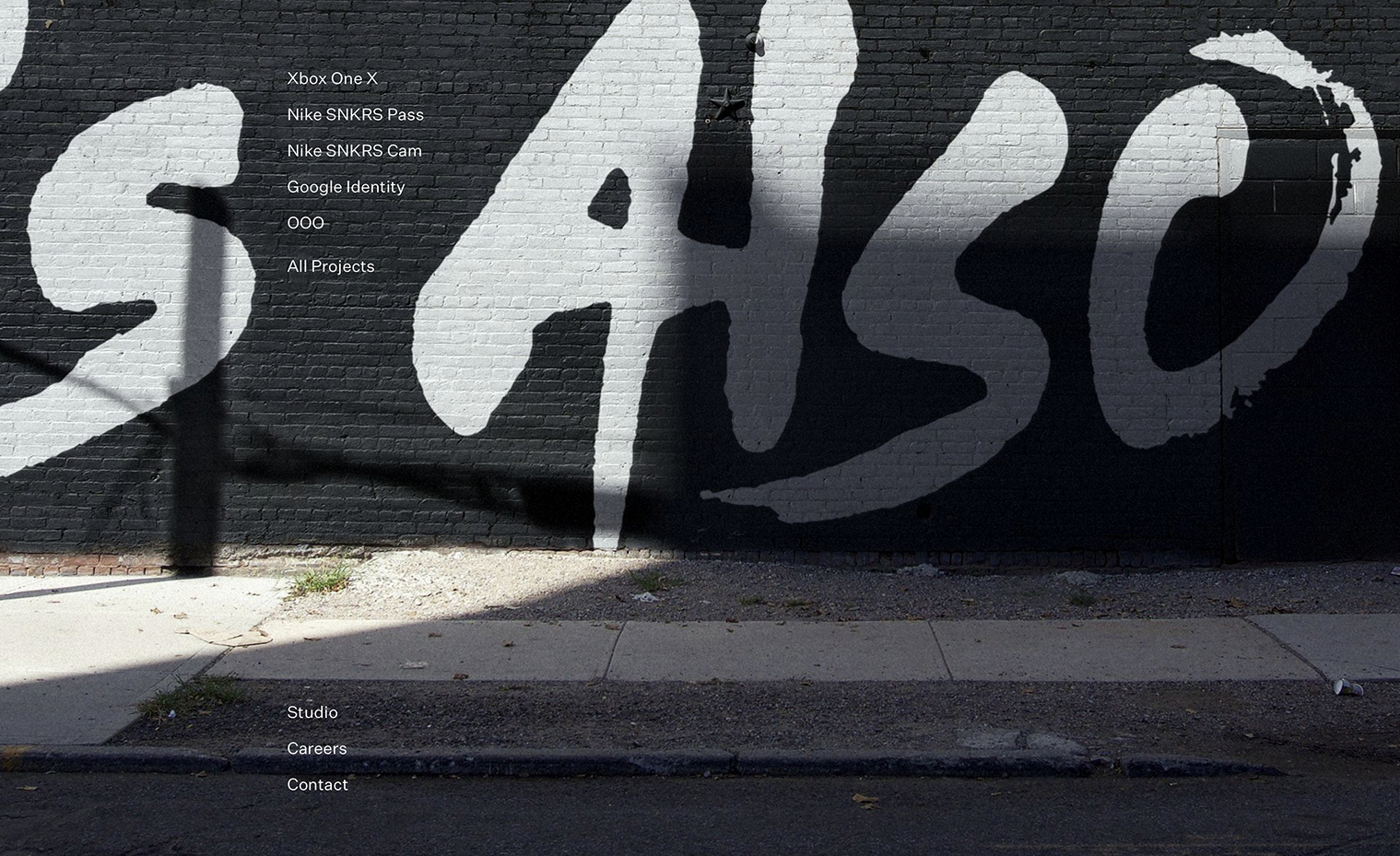 A simple site menu over a full-bleed background image of the This Also logo painted in white over black on the side of a brick building.