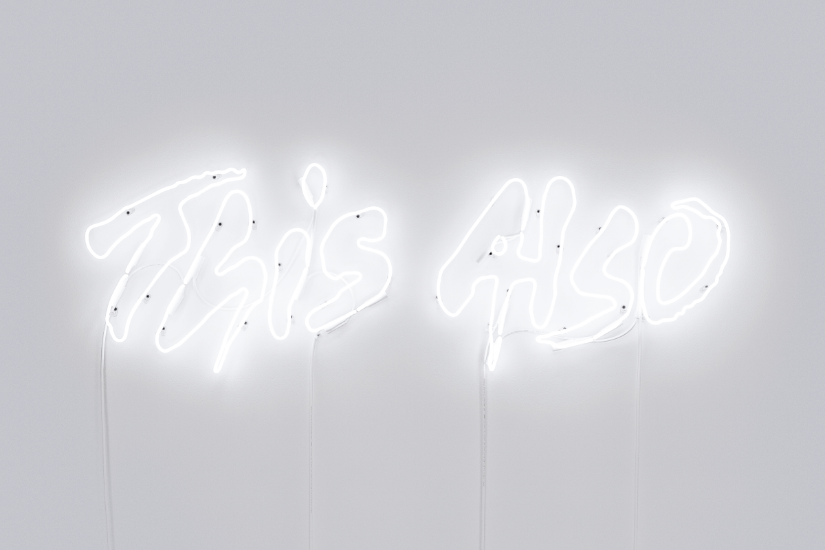 A lit up neon sign of the This Also logo on a light gray wall.